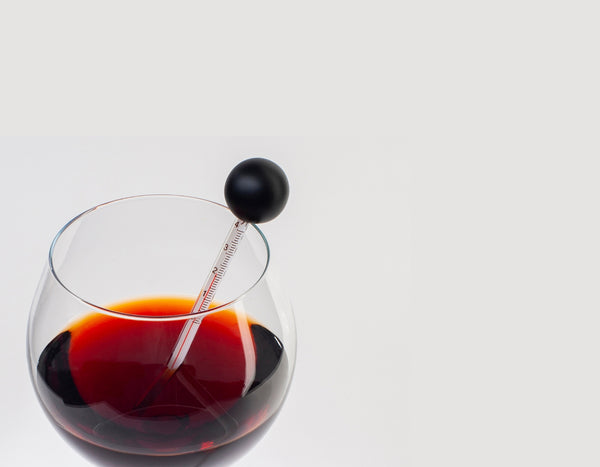 A stemless wine glass with red wine and a glass thermometer; white background.