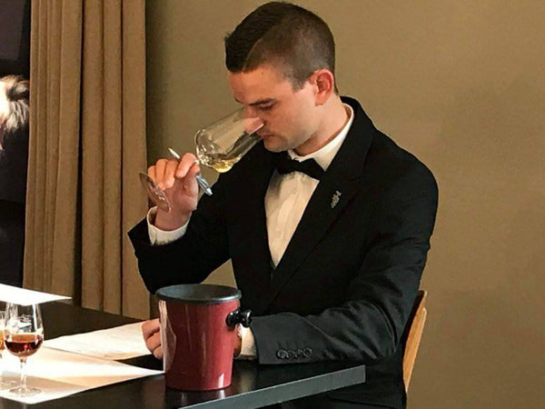 Sommelier in tuxedo tasting wine, indoor setting, with focus on the wine glass and the person's expression.